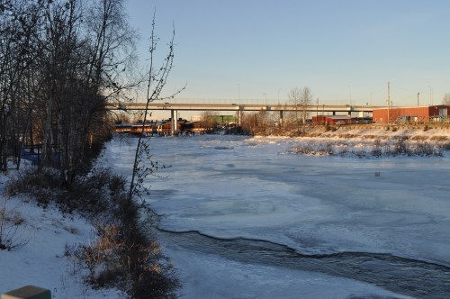 A frozen river scene with snow-covered banks and a bridge in the background under a clear blue sky.