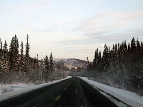 A snow-covered road stretches through a winter landscape with tall evergreen trees and distant mountains under a pale sky.