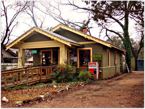 A charming green house with a porch, surrounded by trees and a gravel path, featuring a "For Sale" sign in front.