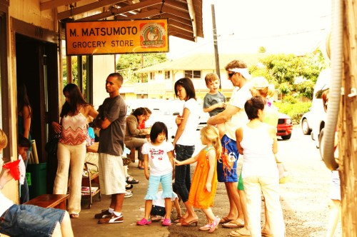 A diverse group of people, including children, waiting outside M. Matsumoto Grocery Store on a sunny day.