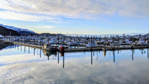 A serene marina with numerous boats docked, reflecting in calm water under a cloudy sky. Snow-capped mountains in the background.