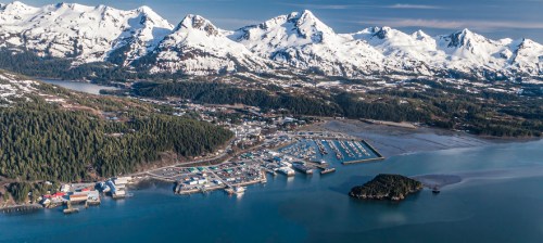 Aerial view of a coastal town surrounded by snow-capped mountains and a marina, with lush greenery and calm waters.