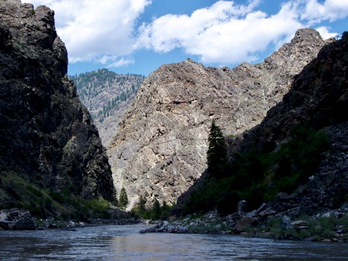 A serene river flows between steep, rocky cliffs under a blue sky with fluffy clouds. Lush greenery lines the banks.