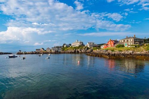 Coastal scene with boats in the water, rocky shore, and charming buildings under a blue sky with clouds.
