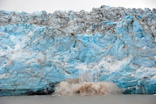 A massive glacier calving into the water, creating a splash and ice debris against a backdrop of blue ice.