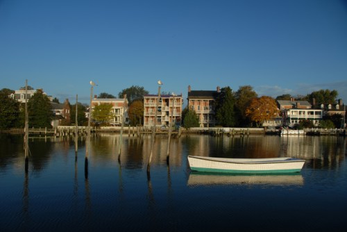 A calm waterfront scene with a small boat and colorful buildings reflected in the water under a clear blue sky.