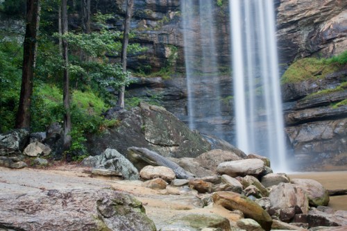 A serene waterfall cascades over rocky cliffs, surrounded by lush greenery and large boulders at the base.