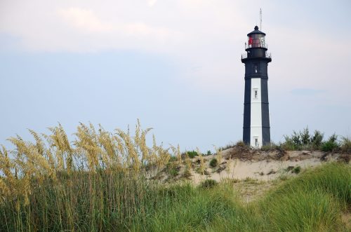 A tall black and white lighthouse stands on a sandy dune, surrounded by tall grass under a cloudy sky.