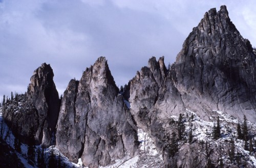 Rocky mountain peaks rise against a cloudy sky, with patches of snow and trees at their base.
