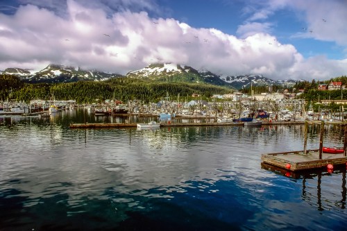 A scenic harbor with boats, surrounded by mountains and lush greenery under a partly cloudy sky.