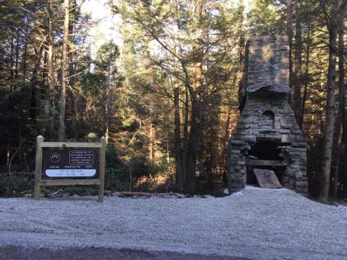 A stone chimney stands among trees, with a sign nearby indicating a trail or park entrance. Gravel path in foreground.