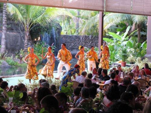 A lively dance performance with performers in colorful outfits, surrounded by tropical plants and an audience enjoying the show.