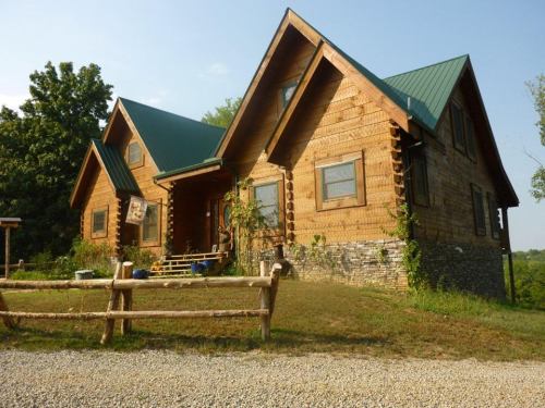 A rustic wooden house with a green metal roof, surrounded by trees and a gravel path.