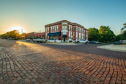 Historic brick street at sunset, featuring a charming building and parked vehicles in a small town setting.