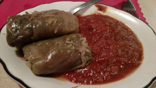 Two stuffed cabbage rolls on a plate with a side of tomato sauce. Fork and spoon are nearby.