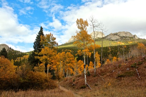 A scenic landscape featuring vibrant autumn trees, mountains, and a clear blue sky with scattered clouds.