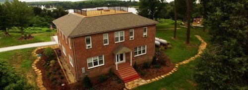 Aerial view of a two-story brick house with a landscaped yard and a rooftop deck surrounded by trees.