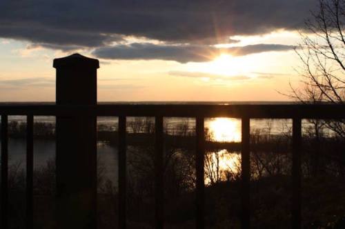 Sunset over a river, with a silhouette of a railing in the foreground and clouds in the sky.