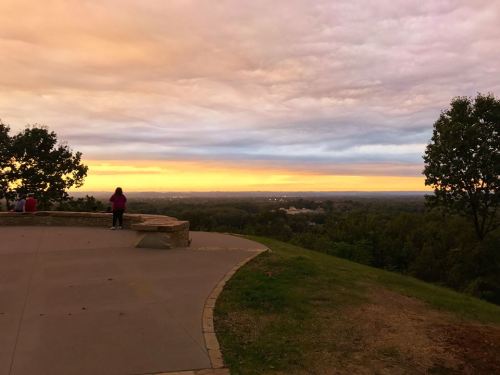 A scenic view from a hilltop at sunset, with people enjoying the landscape and colorful clouds in the sky.