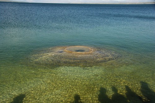 A circular rock formation rises from clear water, surrounded by a calm lake and distant mountains. Shadows of onlookers visible.
