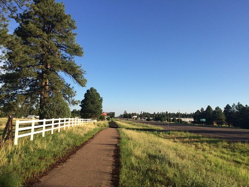 A paved path lined with grass and trees, leading towards a highway under a clear blue sky.