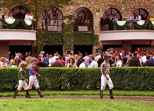 Jockeys in colorful silks walk past a crowd at a horse racing event, with spectators on a balcony above.