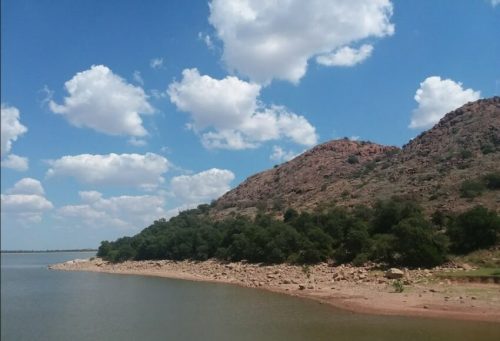 A serene landscape featuring a rocky hill, lush greenery, and a calm body of water under a blue sky with fluffy clouds.