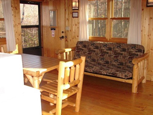 Cozy wooden interior with a table, chairs, and a sofa, surrounded by large windows letting in natural light.