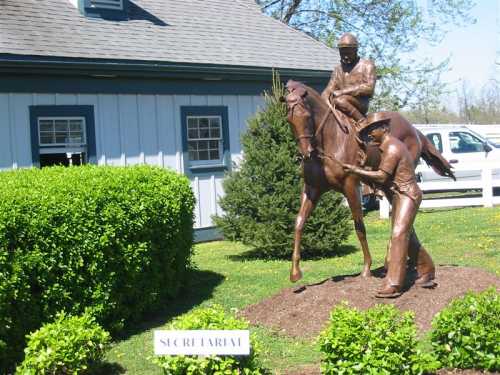 Bronze statue of a jockey on a horse, with a groom holding the horse, set in a landscaped area near a building.