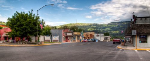 A small town street with shops, trees, and mountains in the background under a partly cloudy sky.