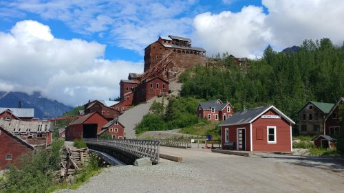 Historic mining buildings on a hillside, surrounded by trees and mountains under a blue sky with fluffy clouds.