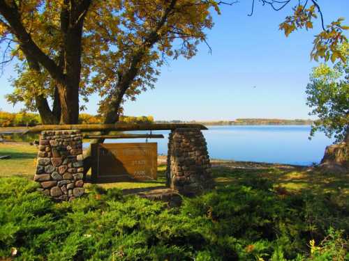 A stone sign for a state park surrounded by trees and a calm lake under a clear blue sky.