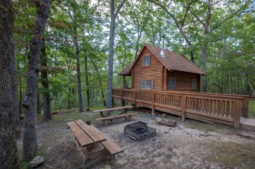 A cozy wooden cabin surrounded by trees, featuring a deck, picnic table, and a fire pit in a serene forest setting.