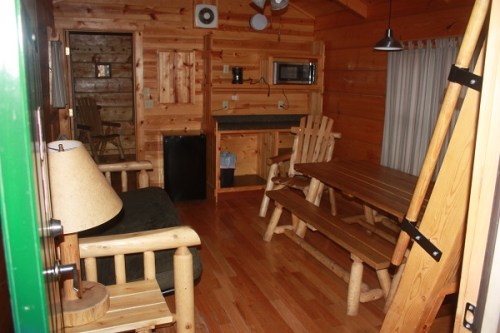 Cozy wooden cabin interior with a sofa, dining table, kitchenette, and warm wooden decor.