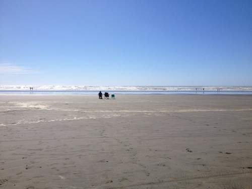 Two people sit on chairs on a sandy beach, with gentle waves and a clear blue sky in the background.