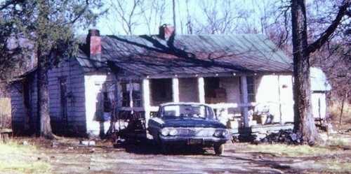 A weathered house with a green metal roof and a vintage car parked in front, surrounded by trees and overgrown grass.