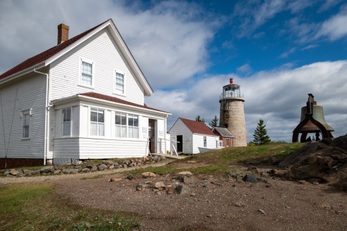 A white house and a stone lighthouse under a cloudy sky, with a sculpture nearby and rocky terrain in the foreground.