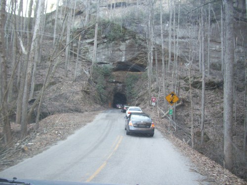 Cars driving through a rocky tunnel on a narrow road, surrounded by bare trees and a caution sign.