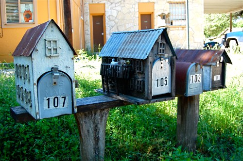 Three unique mailboxes resembling small houses, set on a wooden post in a grassy area.
