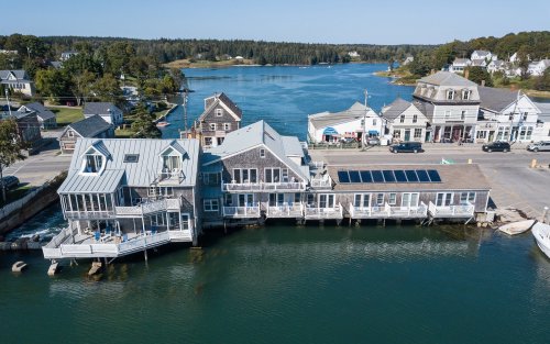 Aerial view of waterfront buildings along a river, with boats and trees in the background on a clear day.