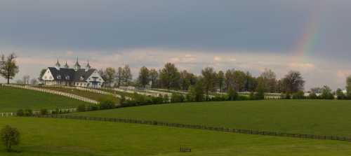 A picturesque landscape featuring a large house with spires, rolling green hills, and a faint rainbow in the sky.