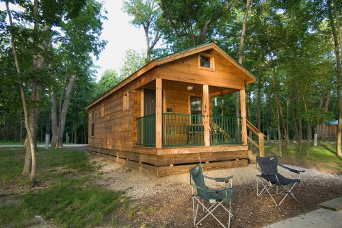 A wooden cabin surrounded by trees, with a porch and two camping chairs in front on a gravel path.