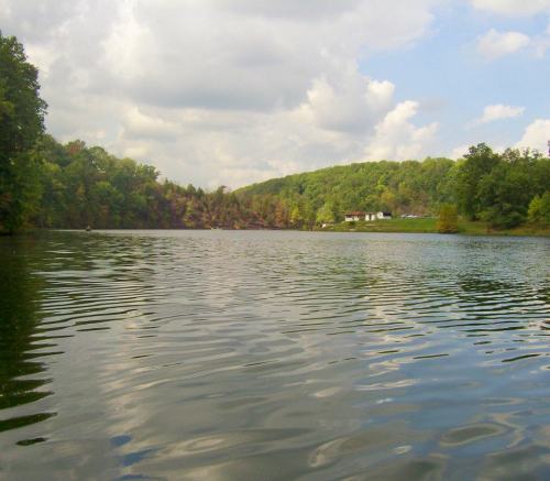 A serene lake surrounded by lush green hills under a partly cloudy sky. Reflections ripple on the water's surface.