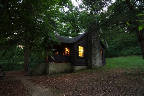 A cozy cabin nestled in a wooded area, with warm light glowing from its windows at dusk.