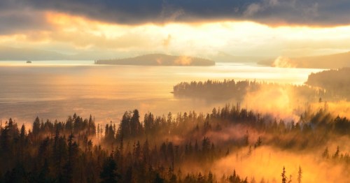 A serene landscape at sunset, featuring a lake surrounded by misty forests and distant islands under a dramatic sky.