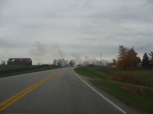A winding road with a cloudy sky, flanked by fields and a distant farm with smoke rising in the background.