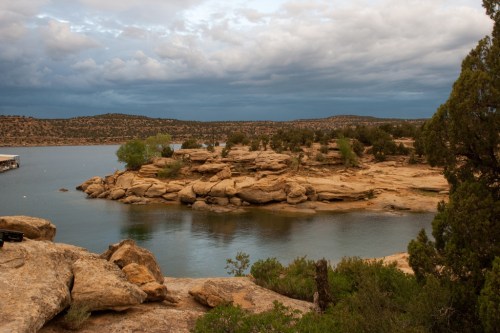 A serene lakeside view with rocky shores, lush greenery, and a cloudy sky in the background.