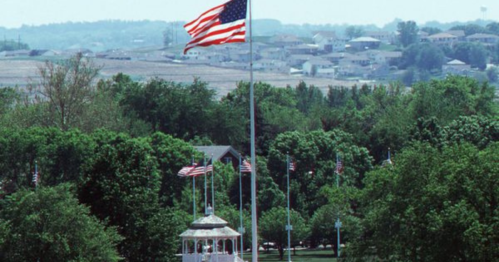 A large American flag waves above a gazebo surrounded by trees and smaller flags, with hills in the background.