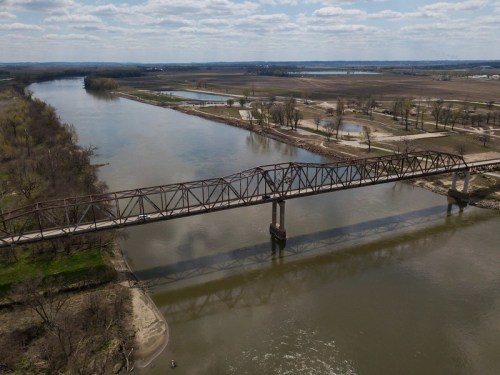 Aerial view of a rusty bridge over a wide river, surrounded by fields and trees under a partly cloudy sky.