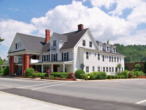 A large white historic building with multiple stories, surrounded by greenery and under a blue sky with clouds.